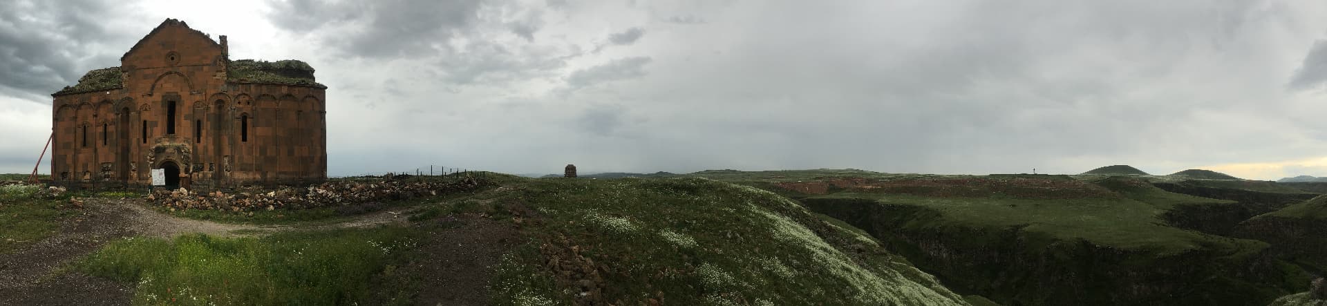 A lone ruined stone church standing on a grassy plateau beneath a heavy sky, a gorge falling away to the right